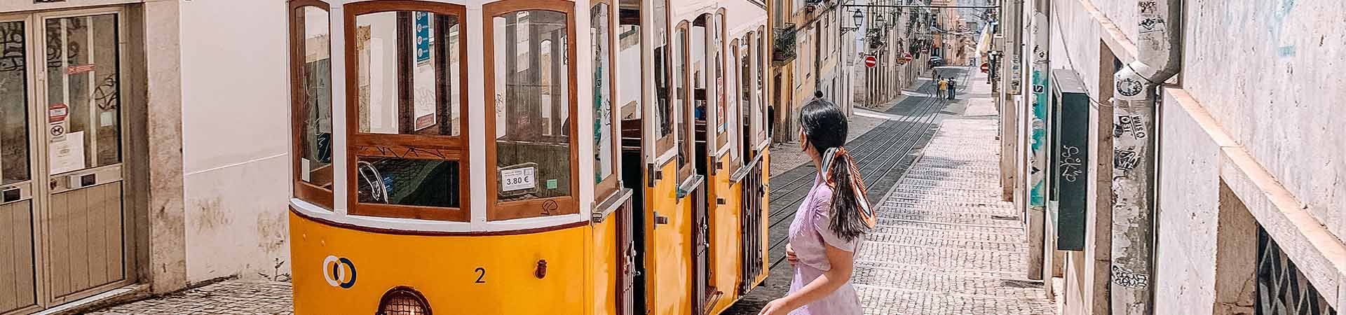 In the center of the image, a yellow and white Lisbon tram passes through one of Lisbon's cobbled streets, lined with buildings with old facades. To the right of the tram, there is a brunette girl, wearing a lilac dress and her hair tied back with a colored scarf, looking at the tram.