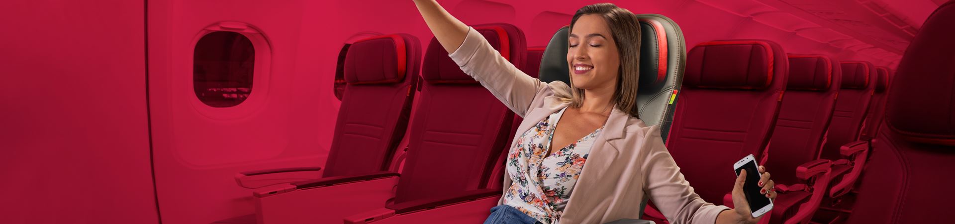 Close shot of a lady sitting in the EconomyXtra seats, inside an airplane. Her eyes are closed, and she is smiling and with open arms. In the left hand she holds a white phone. She is wearing a white sweater with flowers, jeans and a light pink blazer.