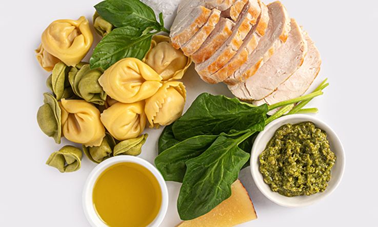Composition with various foods: Below, a small bowl with olive oil, a slice of cheese, some raw spinach and a bowl with pesto sauce. On top, stuffed pasta (yellow or green), two basil leaves and sliced loin meat.