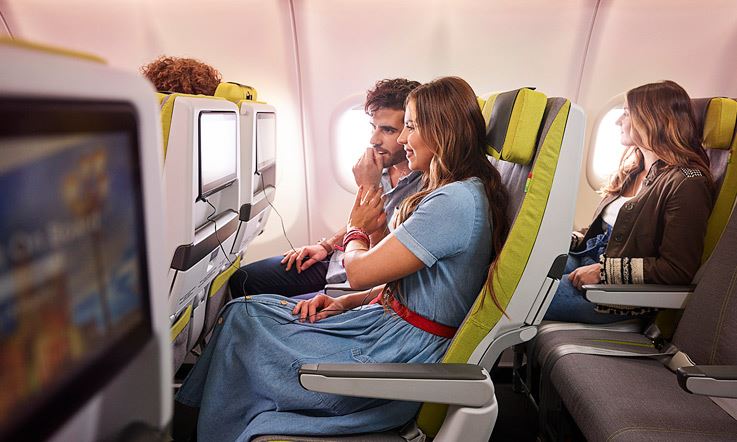 Imagem composta por três passageiros sentados nos lugares verdes de um avião da TAP. Dois dos passageiros estão a olhar para os ecrãs situados nas costas do seu assento da frente. A passageira da fila dde trás está a olha pela janela do avião.